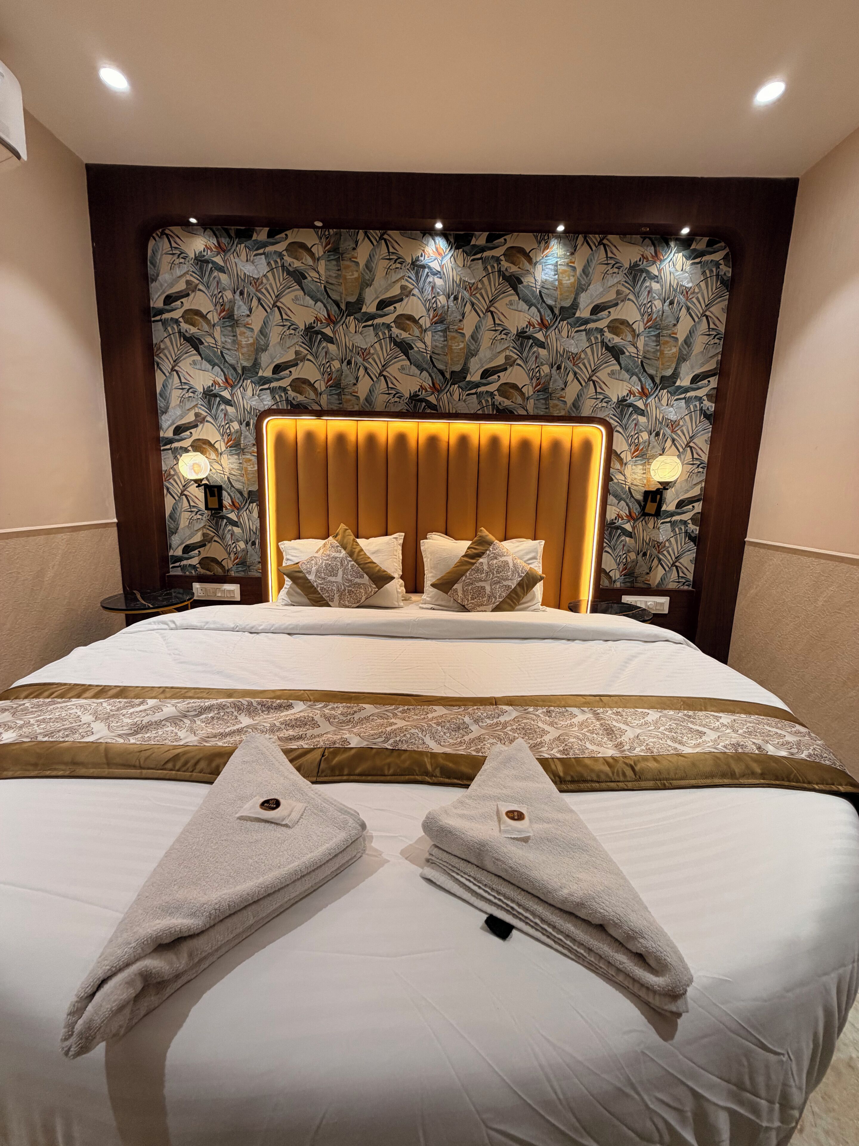 A hotel room with a large bed, a headboard with a floral pattern, and a bedside table with a lamp.