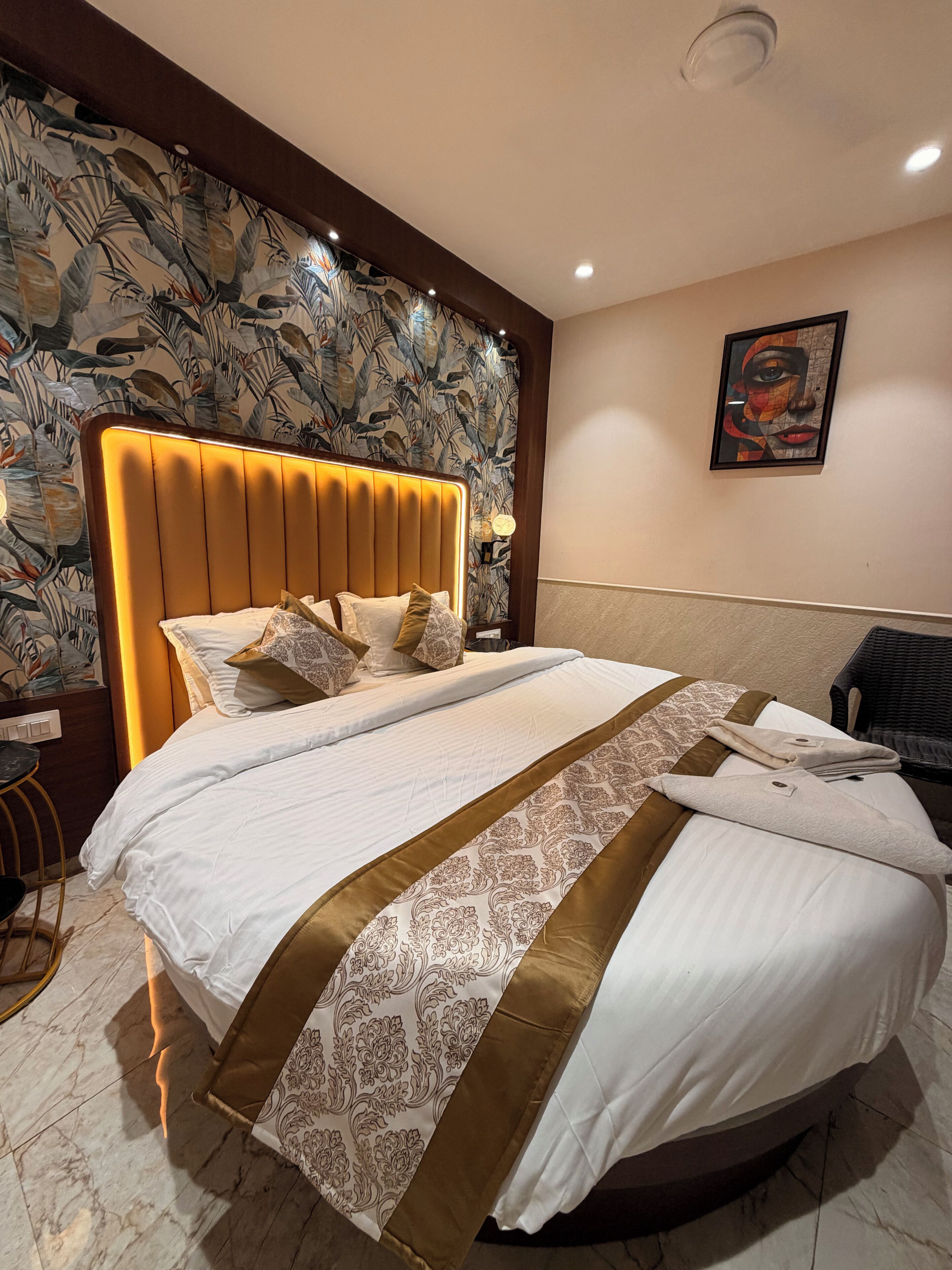 A hotel room with a large bed, a headboard with built-in lighting, a patterned wallpaper, a framed artwork on the wall, and a small table with a chair.