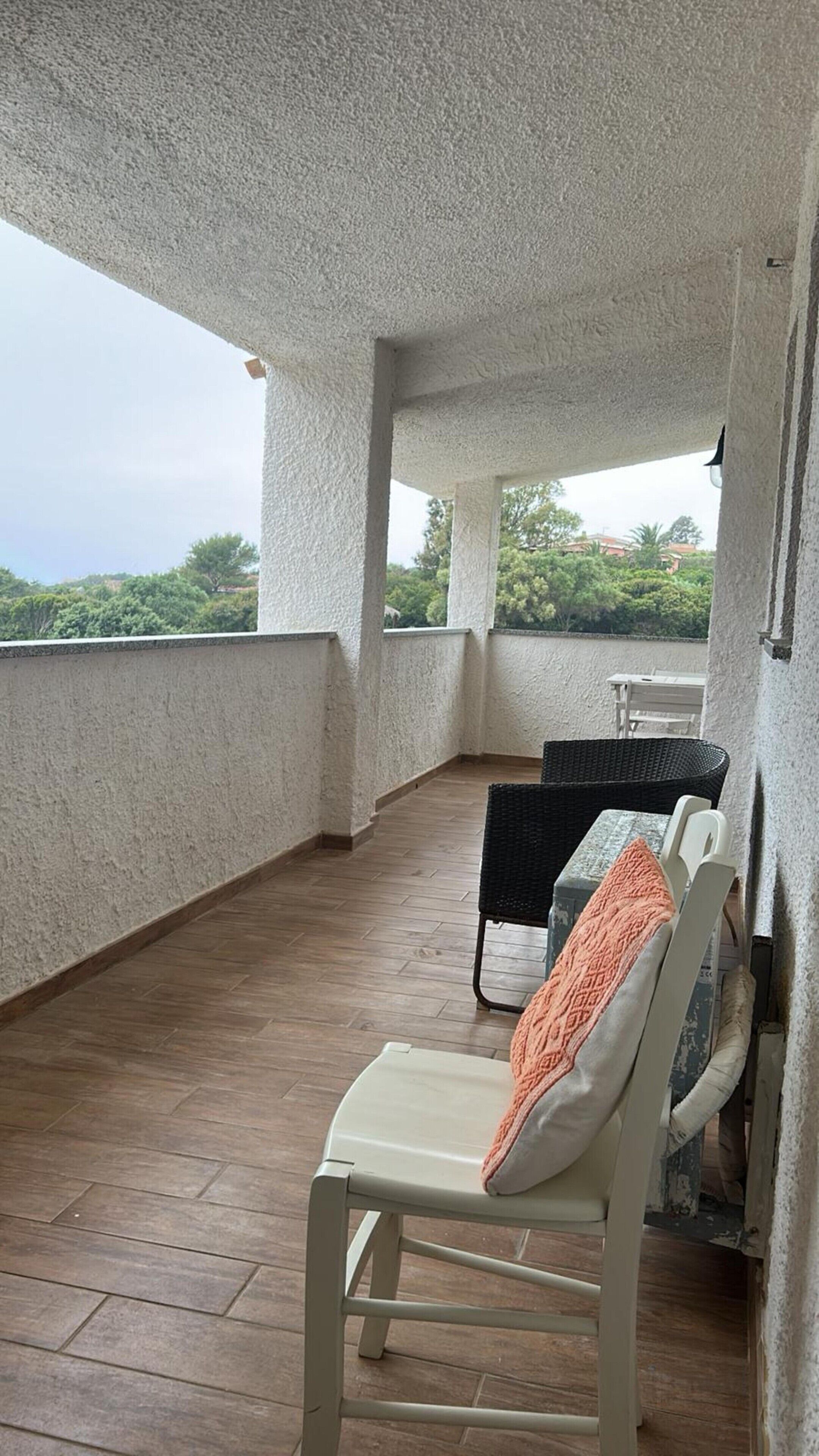 Un balcón techado con suelo de madera, una silla blanca con cojín naranja y una silla de mimbre con cojín blanco.