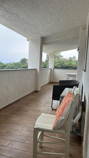 A covered balcony with a wooden floor, a white chair with an orange cushion, and a wicker chair with a white cushion.