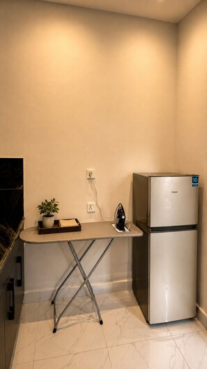 Una cocina compacta con un refrigerador de acero inoxidable, una tabla de planchar pequeña con plancha, una planta en maceta y un enchufe en la pared.