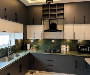 A modern kitchen with dark cabinetry, a gas stove, a coffee maker, and a window. - Spacious 3BR Home in DHA Rahbar - WiFi, Parking & 24/7 Support (Lahore)