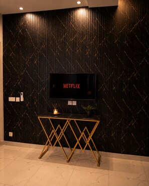 Una habitación moderna con una pared de diseño oscuro, un televisor de pantalla plana montado sobre una consola con marco dorado, y una vela encendida sobre la mesa.