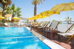 Piscina com espreguiçadeiras e guarda-sóis, cercada por palmeiras e um céu claro.