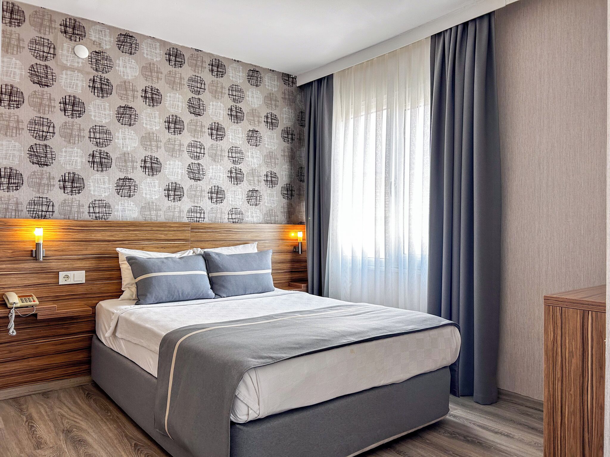 A modern hotel room with a wooden headboard, a large bed with grey bedding, two bedside lamps, and a patterned wallpaper.