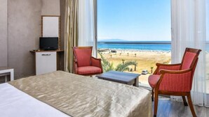 A hotel room with a bed, a TV, two chairs, a small table, and a large window with a beach view. - Acem Hotel (Ayvalik)