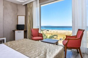 Quarto de hotel com cama, televisão, duas cadeiras, uma mesa pequena e uma janela grande com vista para a praia.