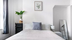Un dormitorio ordenado con cama, mesita de noche, lámpara, espejo y una planta en maceta.