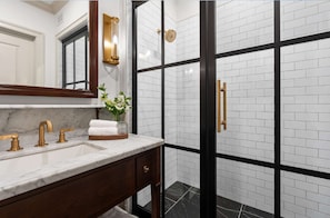 A bathroom with a marble sink, gold fixtures, a mirror, and a black and white tiled shower area. - Historic Leesburg Stay w/ Bunkbeds + Sauna (Leesburg)