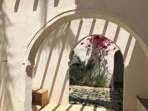 An arched corridor with a tiled floor, a wooden bench, and a view of a garden with plants and a tree.