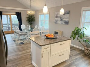 A kitchen with a granite countertop, white cabinetry, and a fruit bowl. There is a dining area with a white table and chairs, and a large window with curtains. - Kate's Catch- 500 Ft To The Beach! (Hatteras)