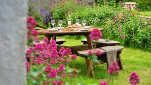 A wooden picnic table with wine glasses and bread, surrounded by lush greenery and flowers.