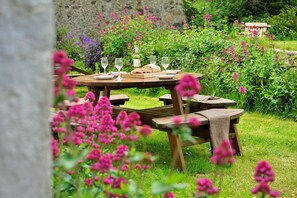 Ein Holztisch für ein Picknick mit Weingläsern und Brot, umgeben von üppigem Grün und Blumen.