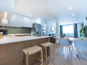 Eine moderne Küche mit einer zentralen Kochinsel, Barhocker, einem Essbereich mit Tisch und Stühlen sowie einem Wohnbereich mit Sofa und Pflanze.