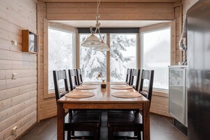 Interior de madera con una mesa de comedor, sillas, una lámpara colgante y vistas a árboles nevados a través de amplios ventanales.