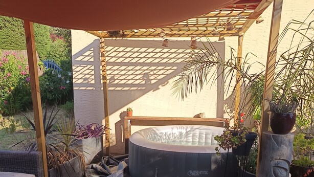 Un patio trasero con jacuzzi, césped artificial, pérgola y plantas en macetas.