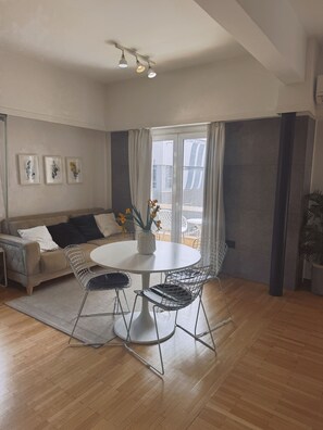 Ein modernes Wohnzimmer mit einer Couch, einem runden Esstisch mit Stühlen, einem großen Fenster mit Vorhängen und Kunstwerken an der Wand.