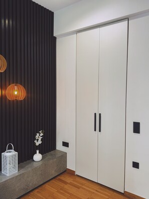 Ein modernes Zimmer mit Holzboden, einer dunkelgrauen Wand mit vertikalen Linien, einem weißen Kleiderschrank mit schwarzen Griffen, einer weißen Vase mit Blumen und einer Pendelleuchte.