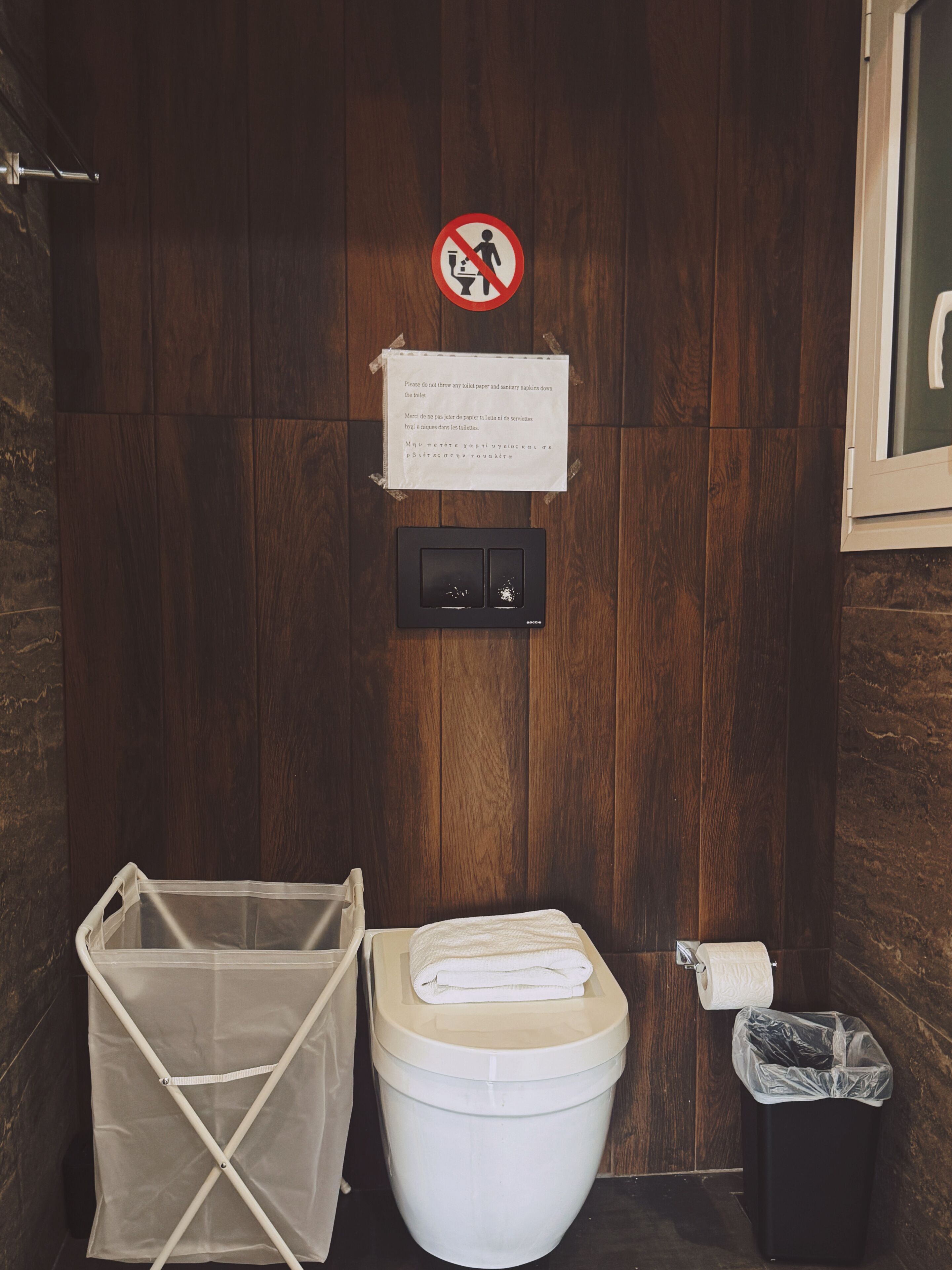 A wooden bathroom with a toilet, a trash bin, and a plastic laundry basket.