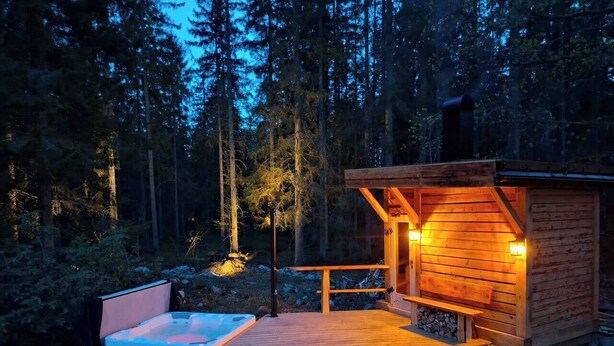 A wooden cabin with a hot tub and a deck, surrounded by a forest.