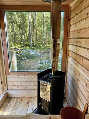 A wooden cabin interior with a large window, a wood-burning stove, and a view of a forest.