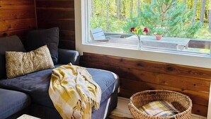 A cozy cabin interior with a sofa, a small table, a wicker basket, and a window with a view of trees.