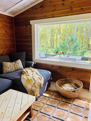 A cozy cabin interior with a sofa, a small table, a wicker basket, and a window with a view of trees.