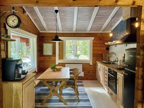 A wooden cabin interior with a kitchen, dining table, chairs, a clock, and a window.