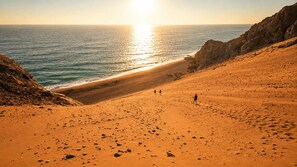 Paisaje desértico con una playa, acantilados rocosos y el sol poniéndose sobre el océano.