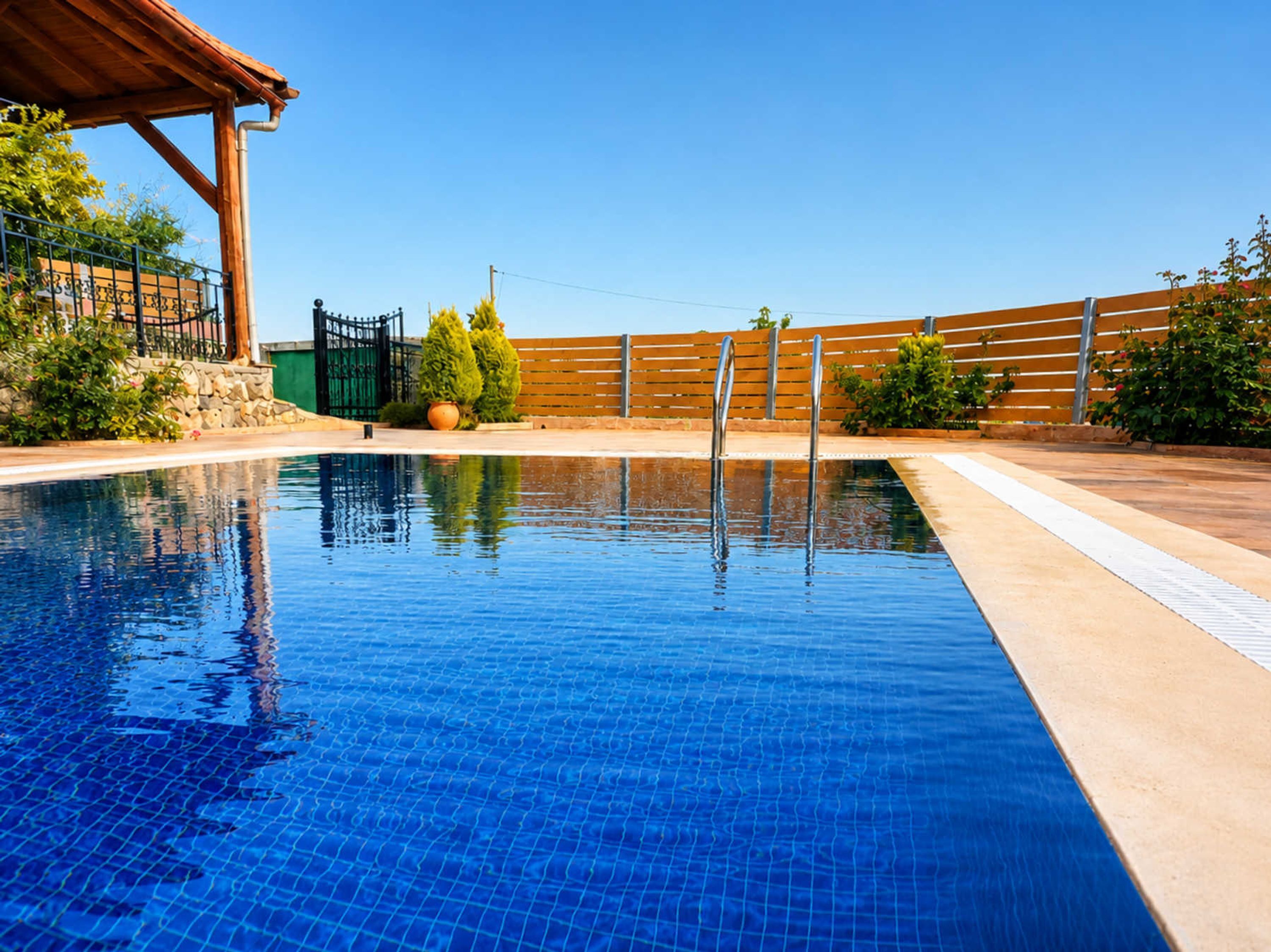 A clear blue pool with a wooden deck, a metal fence, and some greenery.