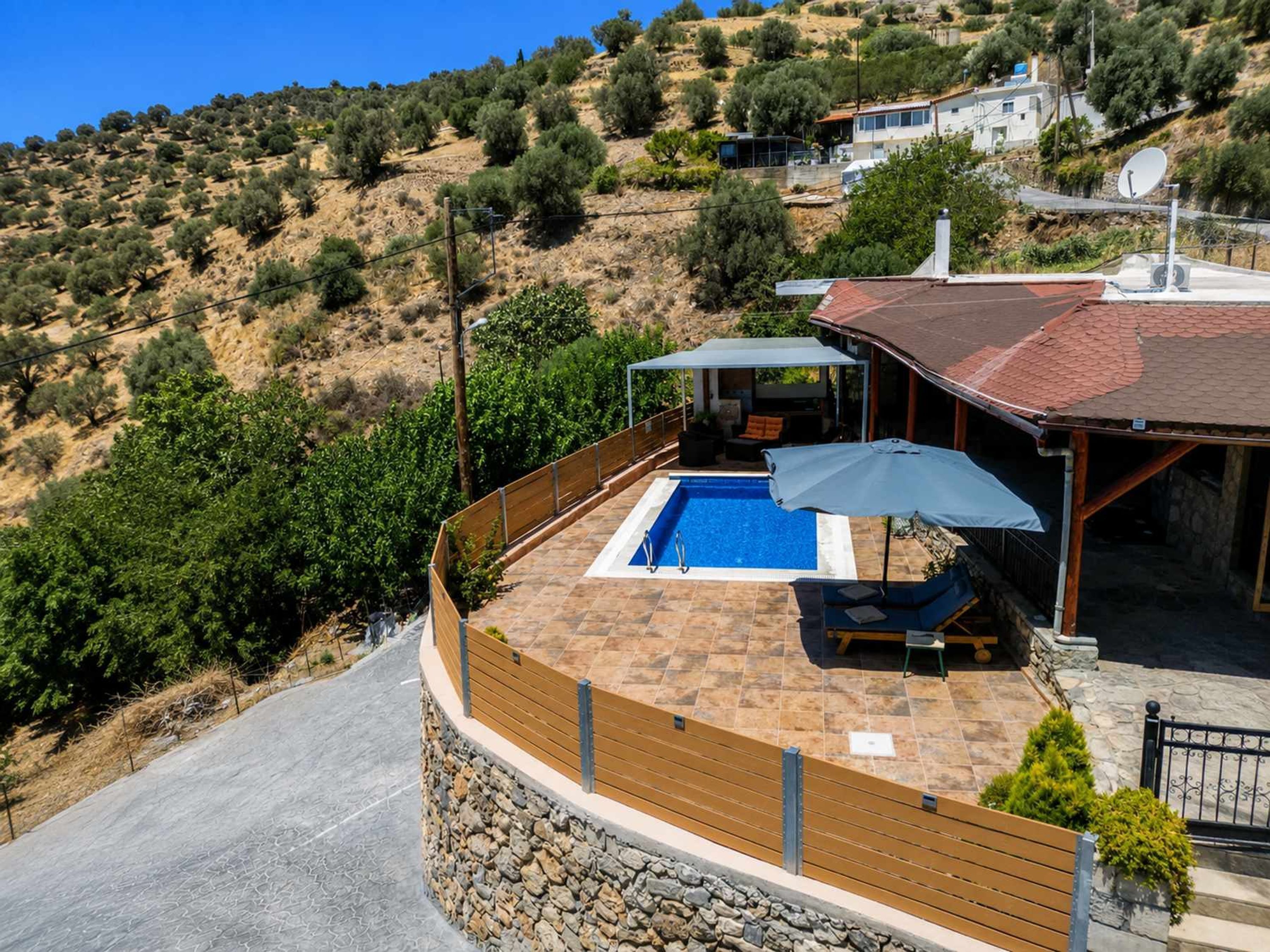 Un área de piscina con un muro de piedra, una terraza de madera y una zona de estar cubierta con mesa y sillas.