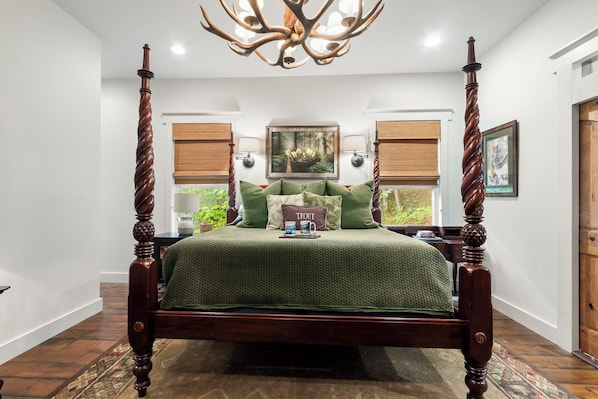 A four-poster bed with a green bedspread, a 'TROUT' pillow, and a chandelier with antler-like arms.