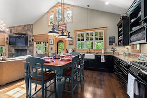 A kitchen with a stone fireplace, a dining area with blue chairs, and a large window with a view of trees. - BEAUTIFUL SETTING - RIVER FRONT W/FIRE PIT! (Murphy)