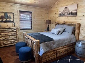 Une chambre à coucher aménagée dans un chalet en rondins de bois, avec un lit en bois, deux poufs, une lampe et un tableau représentant un ours accroché au mur.
