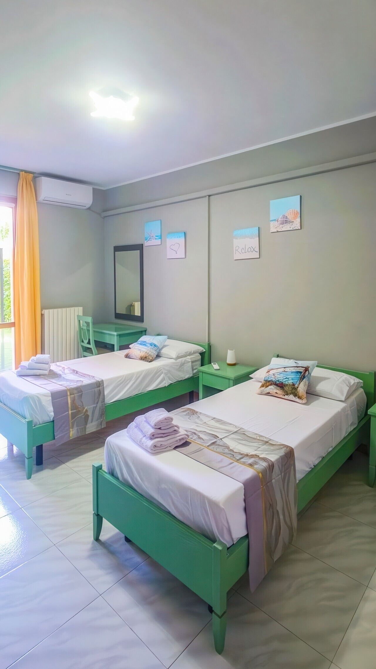 Une chambre d’hôtel avec deux lits verts, une petite table, une chaise, un miroir et une fenêtre avec des rideaux.