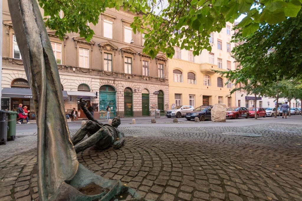 A cobblestone square with a tree, buildings with arched windows, and parked cars.