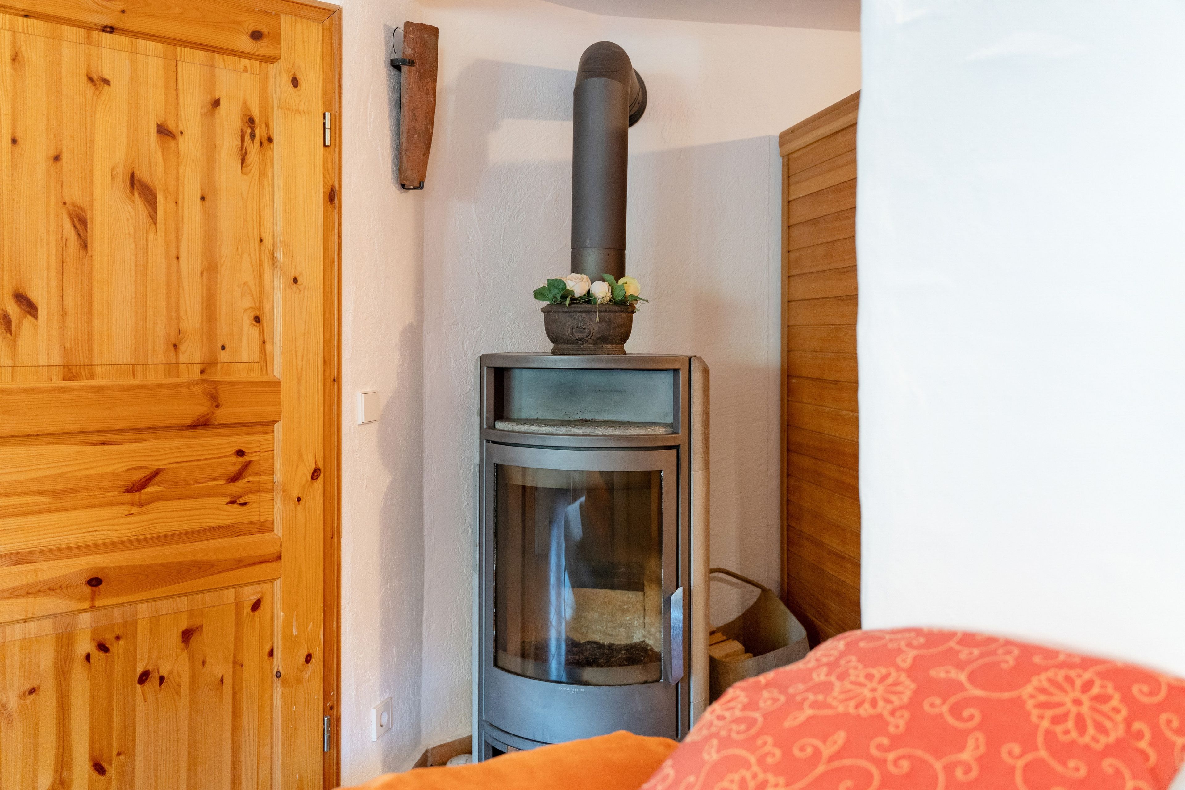 Ein Kaminofen mit einem Blumentopf darauf, eine Holztür und ein gemustertes Kissen.