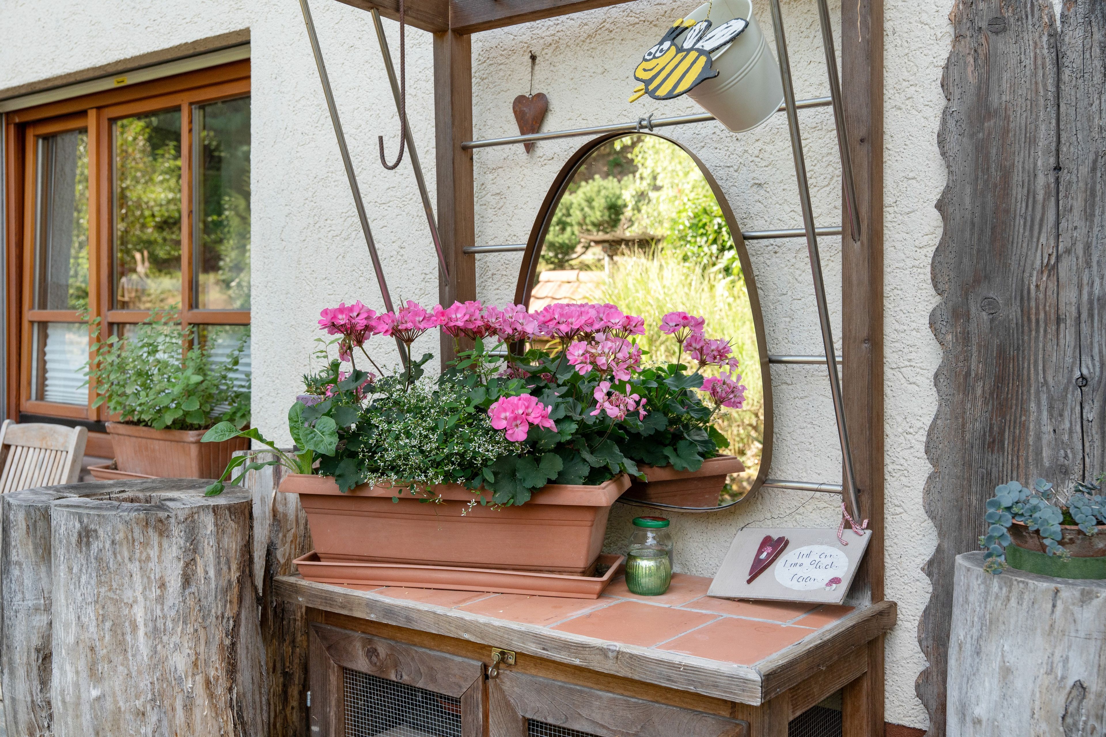 Una ventana con un espejo, una caja de flores con flores rosas y un adorno con forma de abeja.