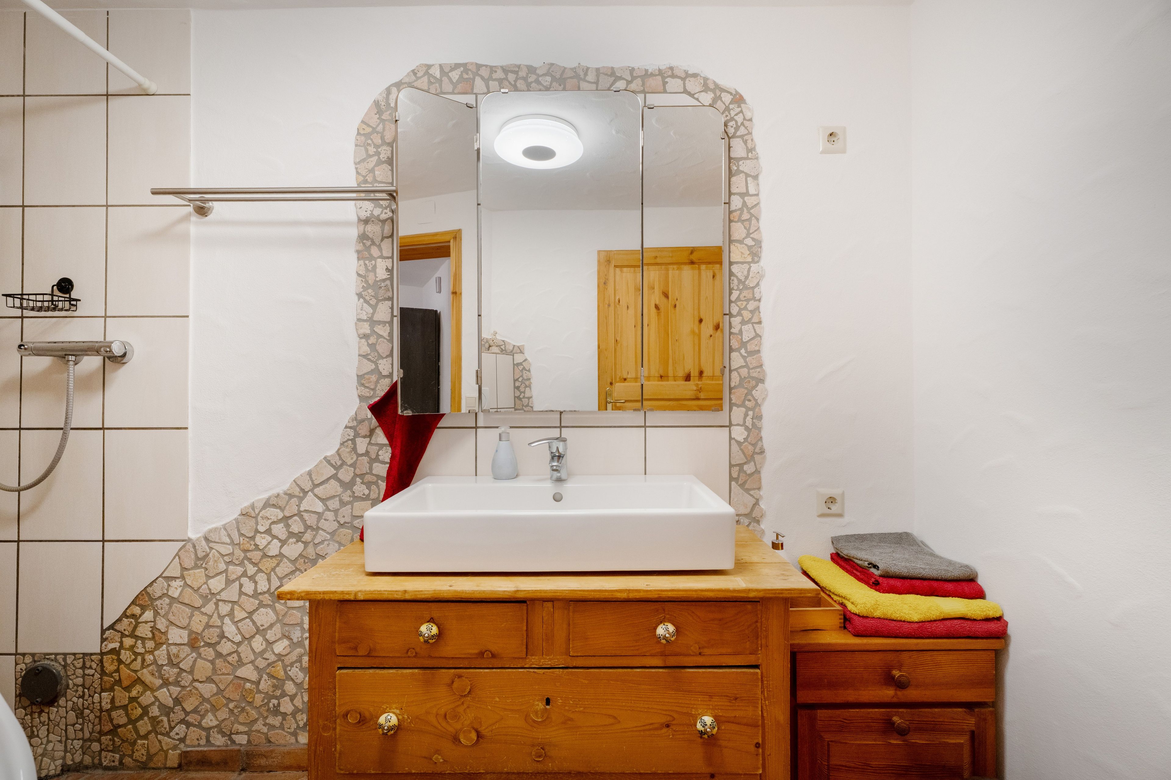 Un baño con lavamanos blanco, tocador de madera, espejo con marco de piedra, zona de ducha y un perchero para toallas.