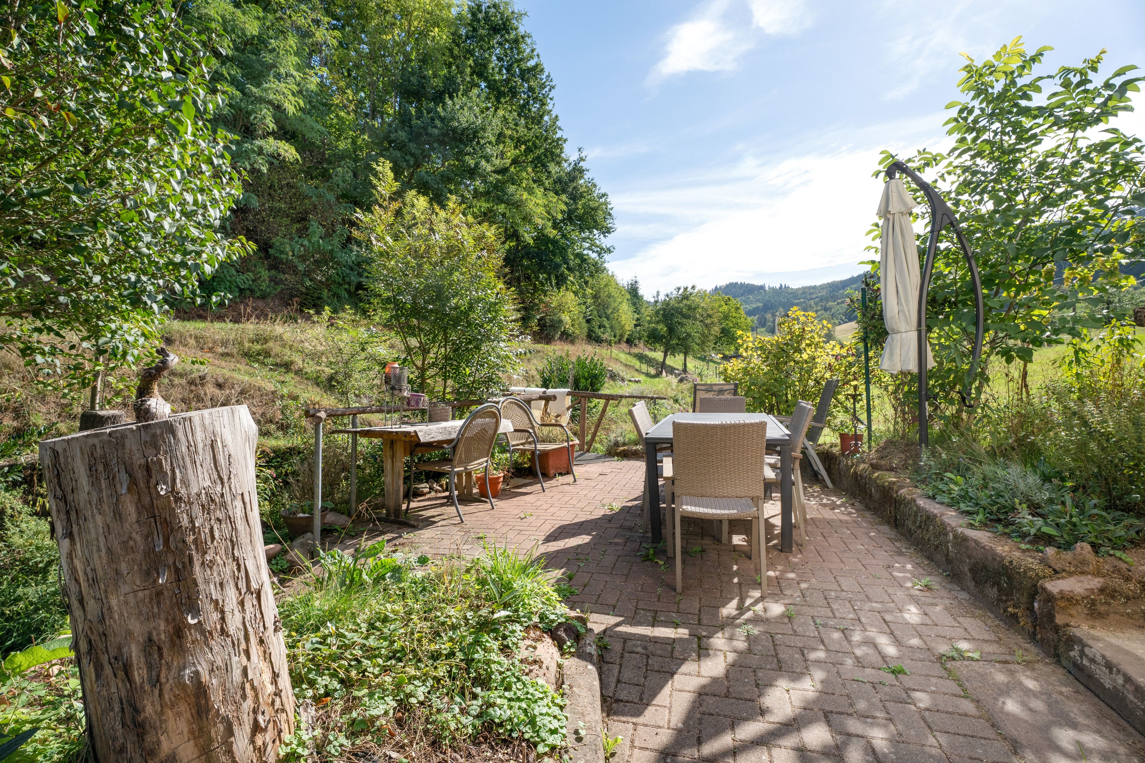 Eine Terrasse im Freien mit Tisch und Stühlen, umgeben von Grünpflanzen und Blick auf das Land.