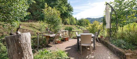 Eine Terrasse im Freien mit Tisch und Stühlen, umgeben von Grünpflanzen und Blick auf das Land.