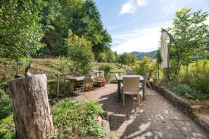 An outdoor patio with a table and chairs, surrounded by greenery and a view of the countryside.