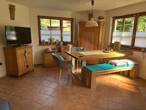 A cozy room with a wooden dining table, a bench, a television on a wooden cabinet, and a window with blinds. - Apartment "Wusslers Bach-Idyll" with Shared Terrace, Garden and Wi-Fi (Gengenbach)
