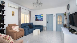 Un salon avec un canapé, un fauteuil et un téléviseur. Il y a un tableau au mur, une porte et une petite table.