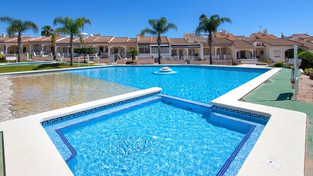 Une grande piscine avec une fontaine, entourée de palmiers et d’un bâtiment doté d’un toit en tuiles.
