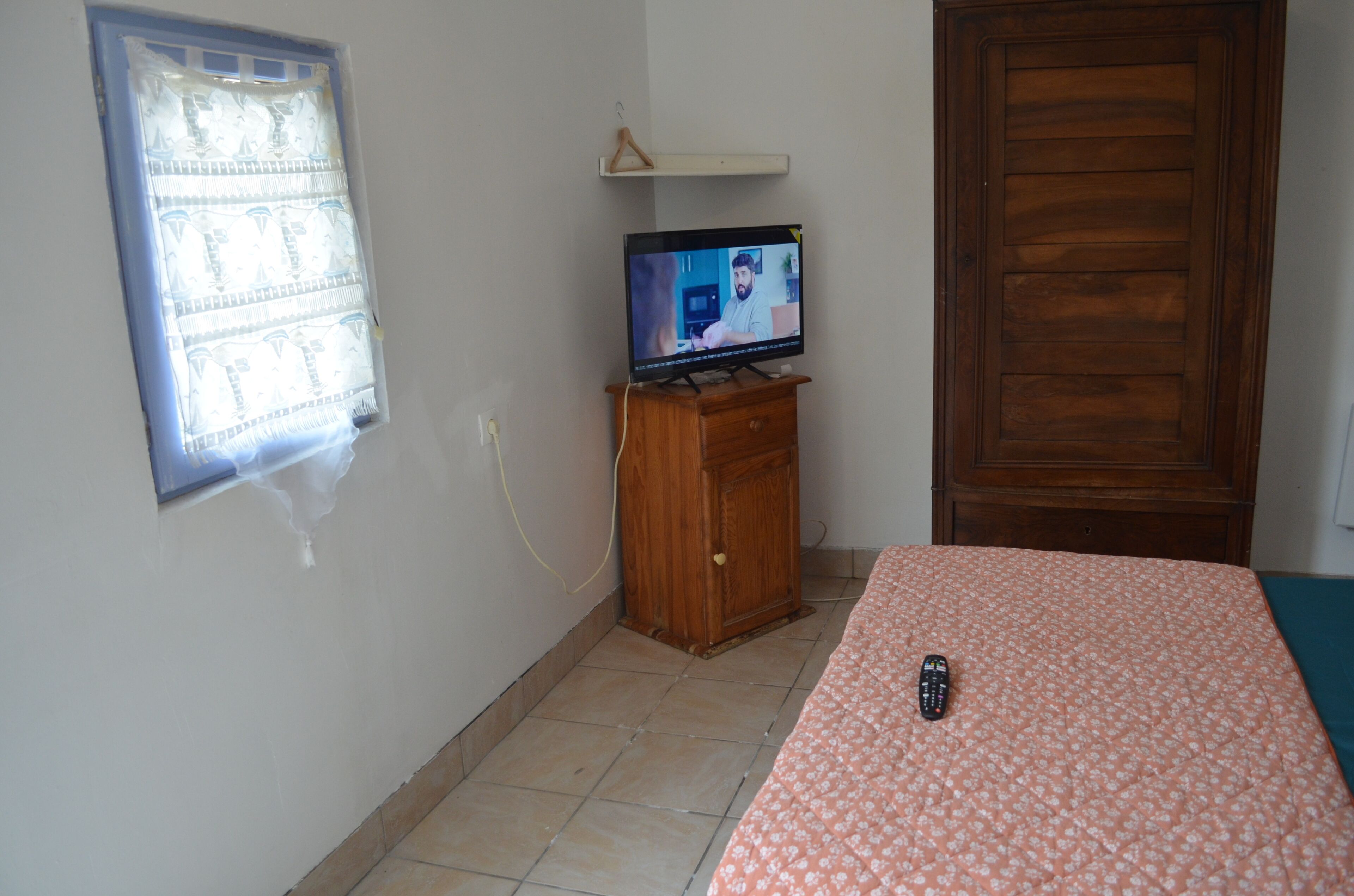 Un dormitorio con una televisión sobre un soporte de madera, una cama con colcha estampada, una ventana con persianas y una puerta de madera.