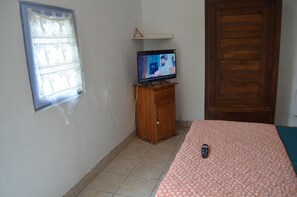 A bedroom with a television on a wooden stand, a bed with a floral bedspread, a window with blinds, and a wooden door.