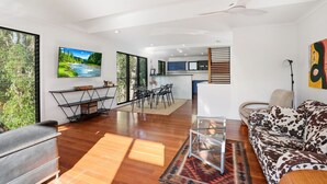 A modern living room with a sofa, a dining area, and a kitchen in the background. - Straddie Beach House 2 | Bush & Beach Retreat (Point Lookout)