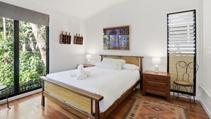 A bedroom with a bed, bedside tables, a nightstand, a chair, a window with blinds, and a wall-mounted picture. - Straddie Beach House 2 | Bush & Beach Retreat (Point Lookout)