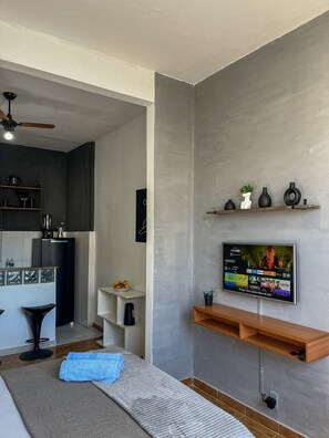 A modern living room with a flat-screen TV, wooden shelves, a ceiling fan, and a kitchen area in the background. - Studio vista mar pertinho da praia (Rio de Janeiro)